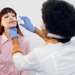 Back view of female afro doctor ent, doing nose examination of her patient. Young Caucasian lady, sitting on the chair and looking at her doctor during nasak check up. Nose deseases concept.