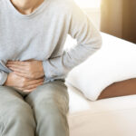 Medical Moment: Understanding PMOS Young woman sitting on a sofa, holding her belly with a painful expression